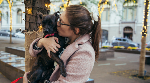 Jamie is holding Lucy in her arms, facing to her right. Her hair is in a bun and she is wearing a light pink jacket. She is kissing the side of Lucy's face. Lucy is wearing a red collar and looking at the camera.
